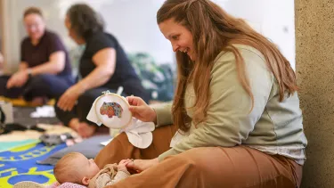 A woman sits smiling on a colorful play mat, engaging with a baby. In the background, other adults and babies interact, creating a warm, joyful scene.