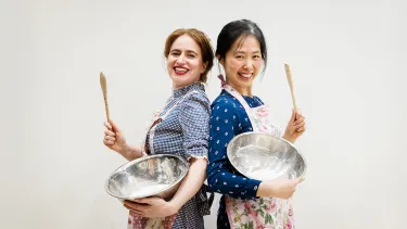 Two smiling women stand back-to-back, holding mixing bowls and spatulas. They wear aprons and convey a cheerful, collaborative mood in a bright setting.