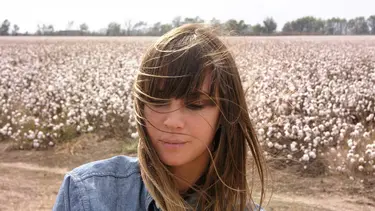 A woman is seen from the shoulders up standing in front of a field of white flowers. She is wearing a blue shirt and has wind swept brown hair.