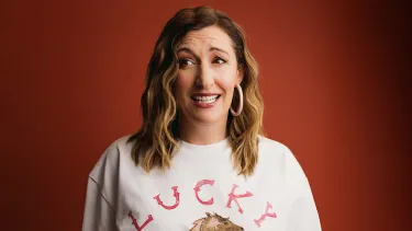 Woman's face sits center, on a maroon background, with an unsure smile and side eyes. She wears a white t-shirt with the word lucky and big hoop earrings as her brown wavy hair falls over her shoulders.