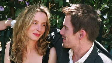A man in a suit jacket and white shirt sits beside a women in a black dress on a green bench. They are looking into each others eyes and the mans arm is around the women resting on the bench.