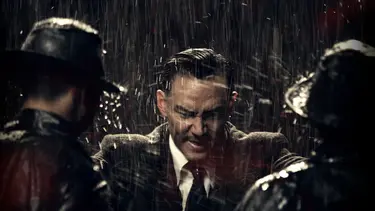 A man in a suit stands in heavy rain, surrounded by two people in dark raincoats and hats, facing him.