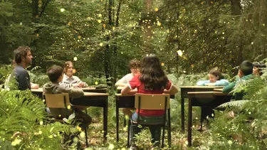 A group of eight people seated around a table in a forest, surrounded by greenery.