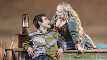 Two actors are seen on the set of the National Theatre Live production The Playboy of the Western World. A man sits leaning back on a desk while a woman leans over him from behind the desk.