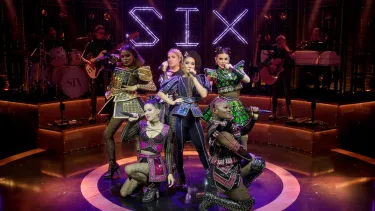 Six performers in colorful, futuristic costumes sing and pose on a stage with a live band behind them and a glowing “SIX” sign above, during a musical performance.