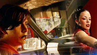 A young man with dark hair inside a car and a woman with a sleek hairstyle in a red dress aiming a firearm.
