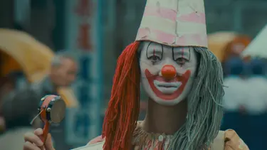 A clown with red and blue hair, and face paint stands in a busy street, holding a tambourine.