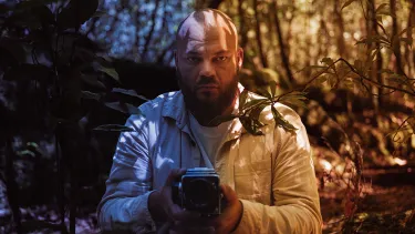 A bearded man is seated amongst trees in a forest. He is holding a retro style video camera.