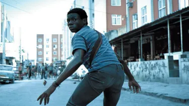 A boy stands semi crouched ready to run in the middle of a street. At the other end of the street there is a group of intimidating figures.