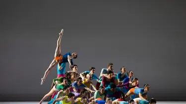 A group of dancers in colorful costumes arranged in various poses on a stage with a neutral background.