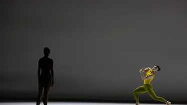 A dancer in a yellow outfit performs a dramatic pose on stage while another dancer in a dark outfit stands in the background.