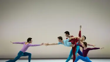 Five ballet dancers on stage, wearing colorful leotards and tights, performing a coordinated group dance with one dancer lifting another.