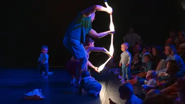 An illuminated performance unfolds in a darkened theatre space as babies and caregivers gather closely around performers manipulating glowing props.