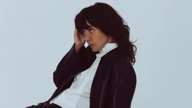 A woman with long brown hair and a fringe sits slumped in a chair slightly side on to the right. They are wearing a buttoned white shirt and black jacket.