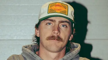 A man wearing a white hat with a green brim and yellow badge stands with his arms crossed. He has a moustache and is wearing a grey hoodie.