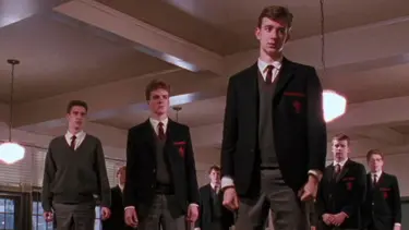 A group of boys in full school uniforms stand on top of desks in a classroom. There are other students still seated.