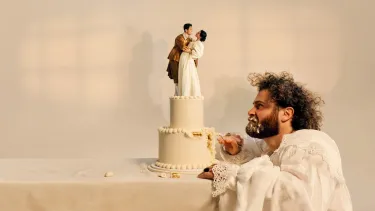 A bearded man picks away and glares at a wedding cake with a happy couple in period costume at the top.
