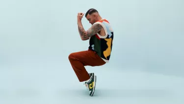 A man with tanned skin and short black hair is posed standing on his toes, with bent knees and his arms held up in front of his face. He wears maroon pants, green and yellow Nike shows and a multi coloured vest.