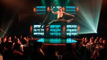 An artist suspended mid-air holding a coat rack in one hand above the stage with an audience gathered around.