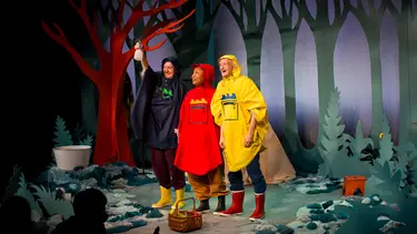 Three performers wearing various coloured rain coats and gumboats stand on a set resembling a forest.