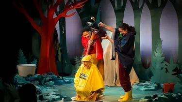 Two performers pour water from watering cans on top of a performer wearing a yellow raincoat.