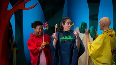 One performer is pointing an electric fan towards two others who are holding up colourful wind spinners.