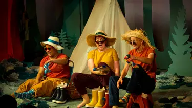 Three performers sit in front of a tent removing their gumboot and socks with amusing looks on their faces. They are wearing an assortment of sunglasses and wide brimmed hats.