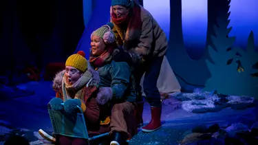 Three performers wearing colourful winter clothes are sitting backwards on a camp chair and miming the act of sledding.