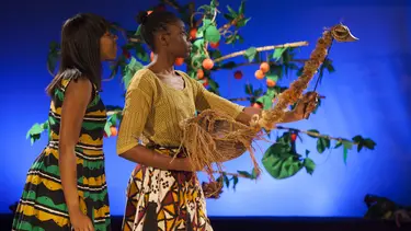 A woman in a colourful green, yellow and brown dress stands next to a performer using an emu puppet. They are in front of a fruit tree and blue back drop.