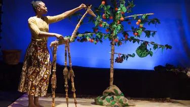 A performer stands in front a tree carrying fruit and a blue backdrop. She is using a puppet of a giraffe.
