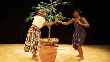 Two women dance around a fruit tree. They are clapping hands together.