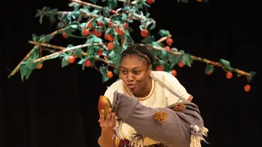 A performer holds a mango in one hand and a puppet that resembles an elephants trunk in the other.