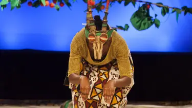 A performer is crouched with her airms out in front while wearing an animal mask and antlers. Behind her is a tree full of fruit.