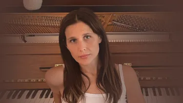 A woman in a white tank top and jeans leans against an upright piano with exposed strings. A white vase sits atop the piano.