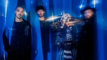 The four members of the band Hiatus Kaiyote stand side by side in dark clothing. They are standing amongst blue streamer like curtains that partially cover them.