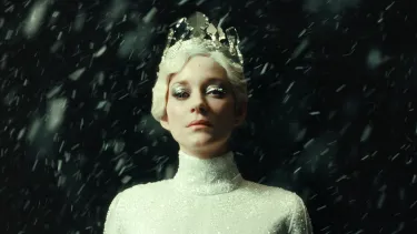 Marion Cotillard stands in a white outfit, with blonde hair and a silver crown. Snow is falling around her with a black backdrop.