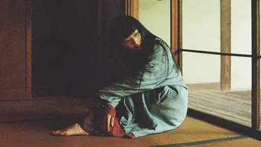 Japanese musician Ichiko Aoba sits on a wooden floor wearing a long faded green garment over the top of a red one that can only be seen near her feet and hands.
