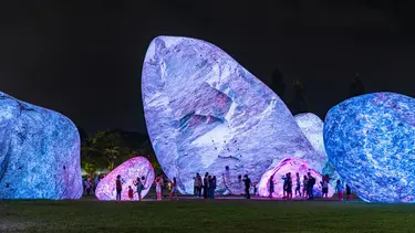 Several large blue and purple glowing rock-like inflatables are in a park. In front of them a group of people walk and take photos.
