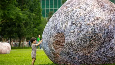 A small child in glasses stands in front of an inflatable rock almost triple his size.