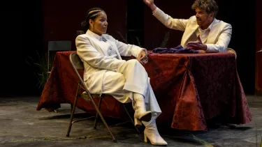 Two actors sit across a desk dressed in white.