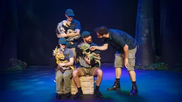Four performers on stage interact with dinosaur puppets, creating a playful, imaginative scene. Dim lighting and forest backdrop add to the theatrical ambiance.