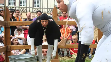 Two performers in sheep suits stand bent over in a pen. They are surrounded by a crowd of adults and children watching on.