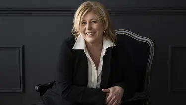 A woman with blonde hair sits in a regal looking black chair. She is wearing a black blazer and trousers and a white shirt with a wide collar.
