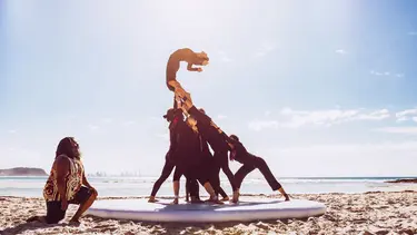 A group of performers in black hold another performer up in the air. They are on a sunny beach with the water behind them. A man in a singlet with a indigenous design is kneeling in front of them.