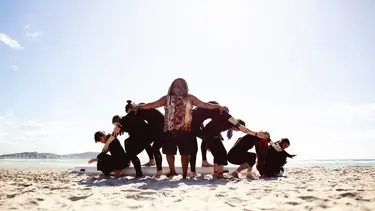 Eight performers are arranged in a symmetrical formation on a sandy beach. At the front is a bearded man wearing a singlet displaying indigenous art.