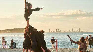 A man in traditional indigenous paint holds a wooden stick up to a group of performers. The performers are holding up a lady doing a ballet pose.