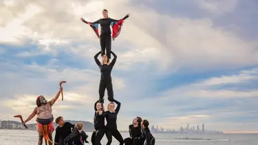 An indigenous man points to the sky where performers have formed a human pyramid standing on each others shoulders. They are on a beach with clouds and a city visible in the background.