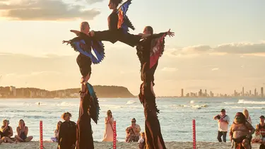 Performers wearing black and coloured fabric resembling wings are on a beach. Two are standing on two others shoulders and they are holding up a fifth performer forming an arch.
