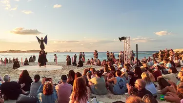 A crowd is seated on a sandy beach watching performers lfiting a person into the air who is wearing fabric wings.