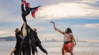A group of performers are holding up a woman in the air. She is posing holding her leg above her head. She is wearing red and blue fabric that resemble wings.
